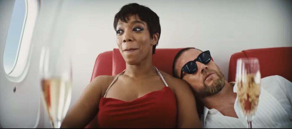 Still from the official trailer for Blink Twice. The image depicts a man with a beard leaning on a young woman in a red dress on an airplane. There are two glasses of champagne in the foreground.